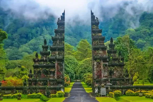Porte monumentale d'un temple balinais, menant à une allée bordée d'arbres luxuriants, avec des montagnes brumeuses en arrière-plan.